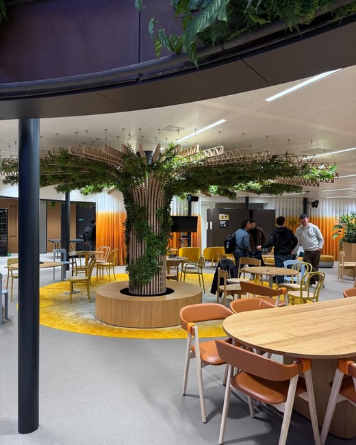 A photograph captures the interior of a modern, brightly colored communal office space featuring a stylized wooden tree structure wrapped in greenery at its center. Several people are gathered near a table in the background, surrounded by modern orange and yellow seating.
