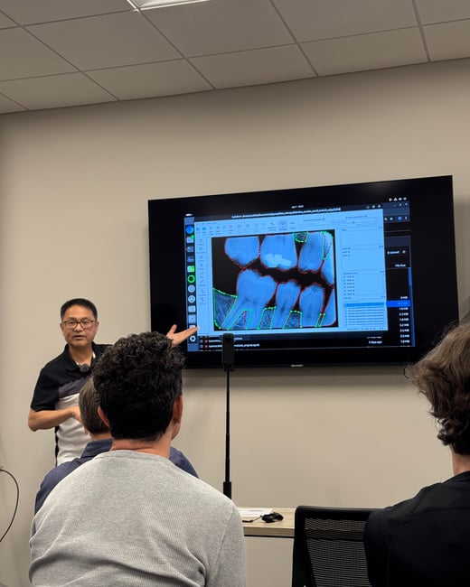 A presenter stands next to a large monitor displaying dental X-ray analysis software, pointing at the screen while addressing an audience.