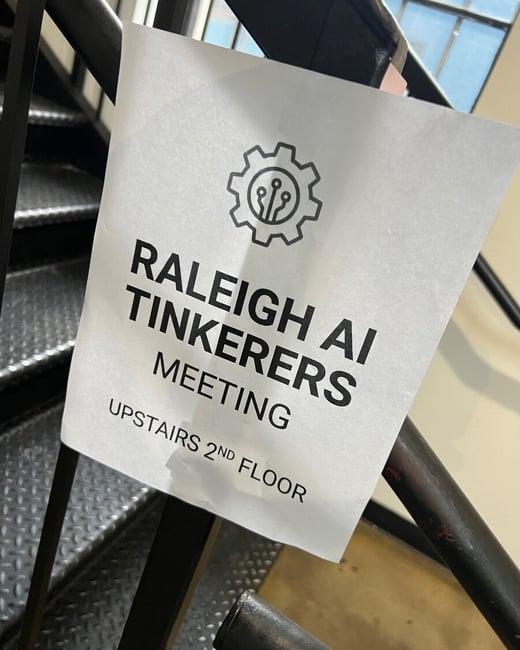 A photograph of a white paper sign taped to a black metal staircase railing, directing attendees to a 'Raleigh AI Tinkerers Meeting' on the second floor.