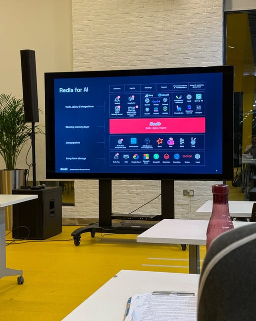 A photograph taken from the perspective of an audience member shows a large monitor displaying a dark-themed technical presentation slide titled "Redis for AI" in a modern indoor space with white brick walls and a bright yellow floor.