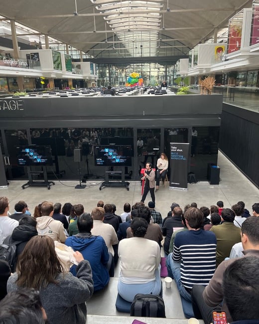 A high-angle photograph captures a large indoor event where a speaker, standing on a stage, addresses a substantial audience seated on tiered steps in a modern, industrial-style atrium, featuring a prominent "Google for Startups" sign in the background.