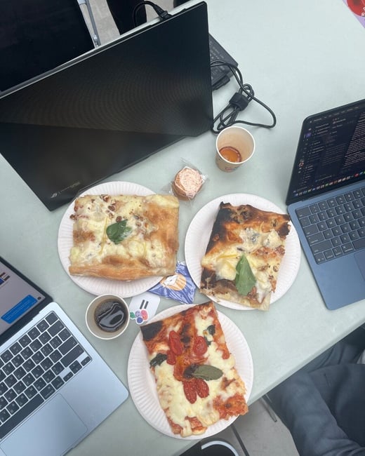 A high-angle photograph shows three slices of rectangular pizza on paper plates, accompanied by two open laptops, small cups of beverages, and a packaged snack on a light-colored table, suggesting a working lunch or study session.
