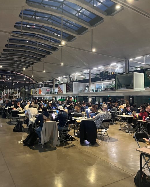 A wide-angle photograph captures a large group of people working intensely on laptops at folding tables in a vast, modern indoor venue, likely during a hackathon or large workshop event.