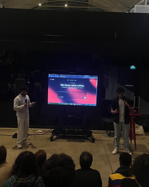 A photograph captures an indoor presentation in a dimly lit auditorium, showing two young men standing in front of a large screen displaying a website with the headline, "We know what works," while an audience sits facing the stage in the foreground.