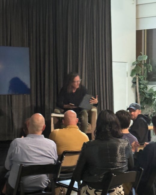 A dimly lit indoor photograph capturing a speaker seated on a stool, holding an object, addressing a small audience who are seated in chairs facing the front. The background consists of dark curtains and a large blue abstract canvas or screen.