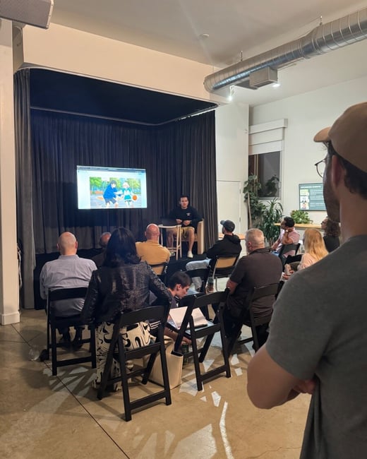 A candid photograph captures an indoor presentation or screening event where a small audience is seated, watching a screen displaying an animation while a speaker sits nearby on a stool. The image is taken from the perspective of someone standing in the back, showing the backs of the attendees.