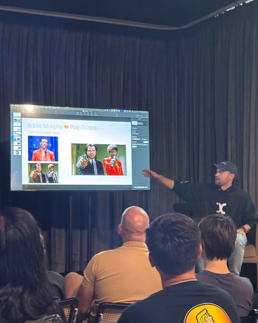 A candid photograph taken during an indoor presentation where a speaker, dressed in a black hoodie, points to a large screen displaying a slide comparing images of Eddie Murphy and characters from Pulp Fiction.