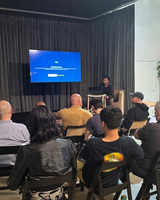 This indoor photo captures an audience seated in an event space, facing a large screen that is displaying a blue error message ("Check Device Power"), with a presenter sitting nearby using a laptop.