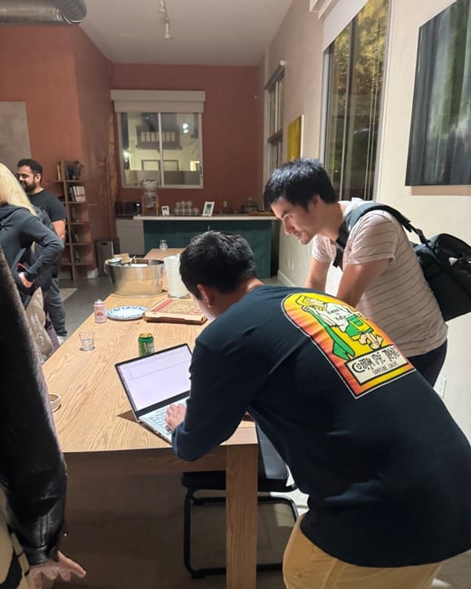 Two men lean over a wooden table to view a laptop screen, collaborating or reviewing data during a casual indoor evening gathering with other people visible in the background.