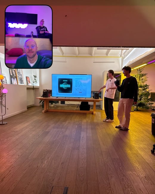 A BeReal photo capturing an indoor presentation scene where two young men stand in front of a TV screen displaying a hackathon slide, juxtaposed with a smaller inset selfie of a third man and another person seated behind him.