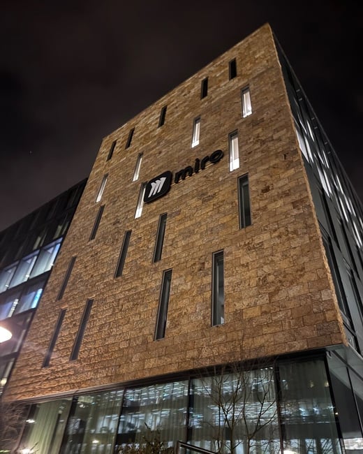 A low-angle nighttime photograph of a modern office building featuring a facade of rough brown stone and numerous vertical windows, with the illuminated "miro" logo prominently displayed near the top.