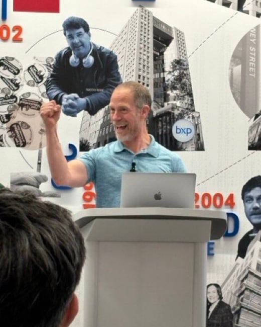 A man in a light blue polo shirt stands behind a podium with a laptop, raising his fist enthusiastically during a presentation in front of a graphic backdrop.