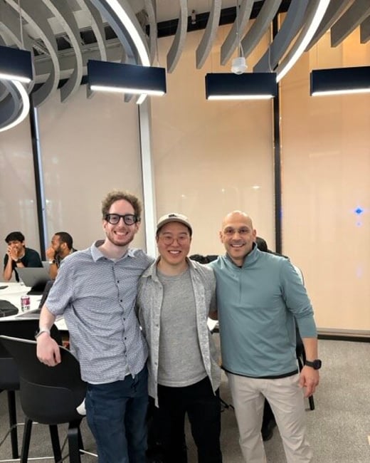 Three men stand close together smiling for a photo in a modern office environment with others seated in the background.