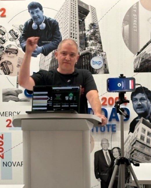 A man stands behind a podium giving a presentation with a laptop open and a smartphone mounted on a tripod nearby.