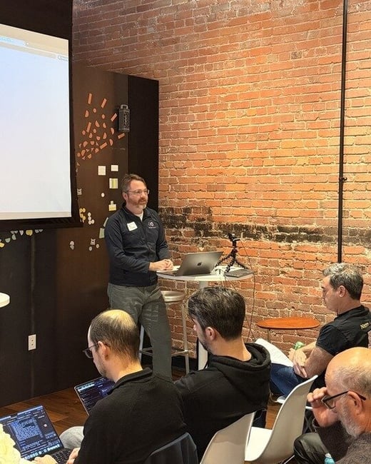 A man stands in front of a projector screen, giving a presentation to a seated audience in a room with an exposed brick wall.