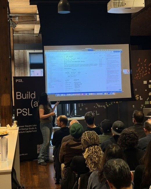 A woman is giving a technical presentation to an audience, with a large screen displaying code or documentation and a 'Build (at) PSL' banner to the left.