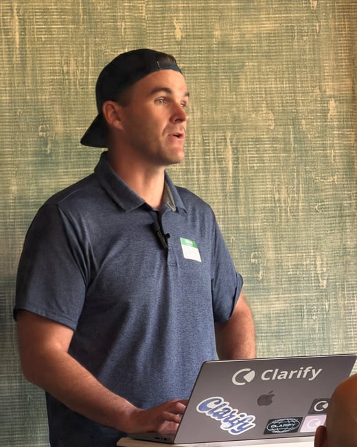 A man wearing a backward baseball cap and a blue polo shirt stands behind a laptop, giving a presentation in front of a textured green wall.