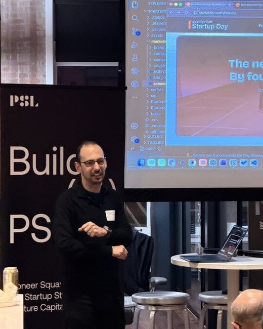 A man in a black sweater and glasses is giving a presentation in an indoor setting, standing next to a PSL branded banner and a projection screen displaying code and a website.