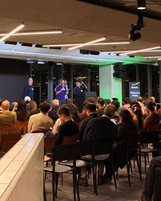 A photograph capturing an indoor corporate or technical event, showing three speakers standing on a stage addressing a large, seated audience in a modern venue with exposed ceilings and green accent lighting. Shopify branding is visible on a screen to the right of the stage.