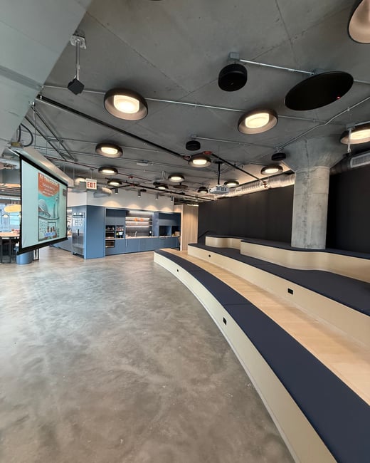 A modern, industrial-style indoor event space featuring tiered wooden seating with blue cushions, a concrete floor and ceiling, and a projector screen displaying an event announcement.