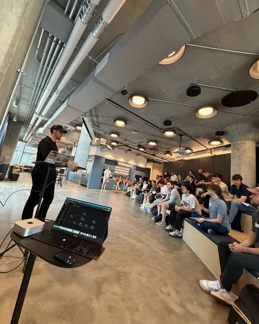 A man is delivering a presentation to a group of people seated on tiered benches in a modern, industrial-style office space.