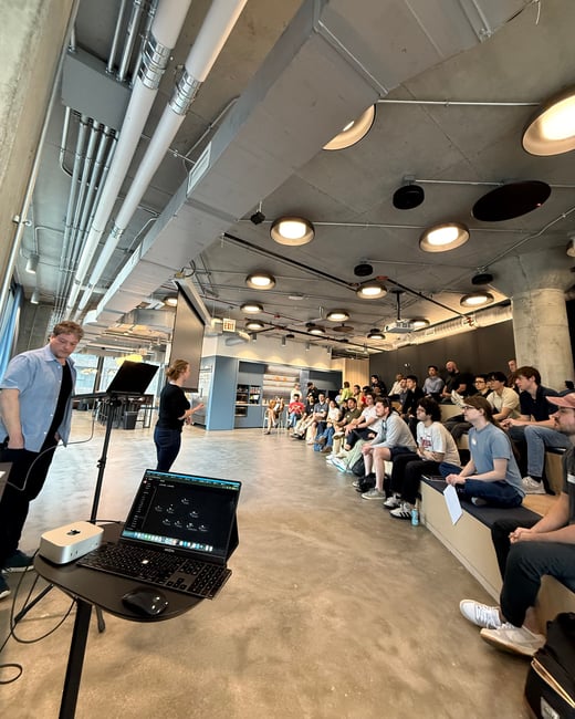 A woman gives a presentation to a seated audience in a modern, industrial-style office space with exposed concrete and piping. A man stands in the foreground near a laptop and monitor setup on a high table.