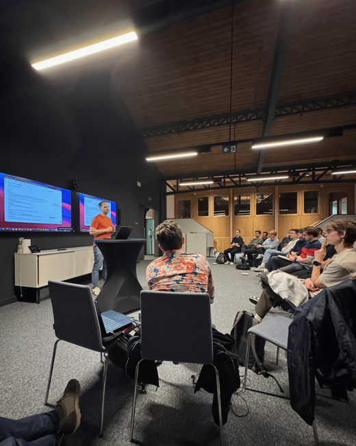 A low-light photograph captures an indoor presentation where a man in an orange shirt speaks to an audience seated in a modern room featuring dark walls, a high wooden ceiling, and two large display monitors showing code or technical content.