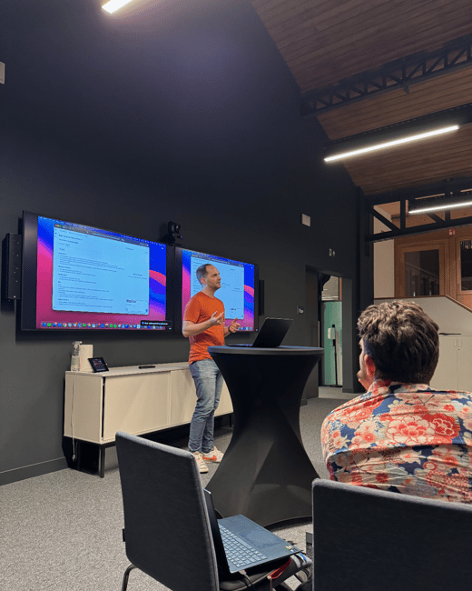 A man in an orange shirt is giving a technical presentation in a modern, dimly lit room, standing between two large wall-mounted screens displaying computer content, while an audience member sits in the foreground with their back to the camera.
