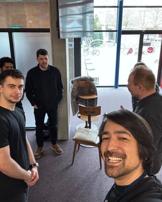 A selfie of a smiling man in the foreground with a group of other men standing in an indoor common area with large windows in the background.