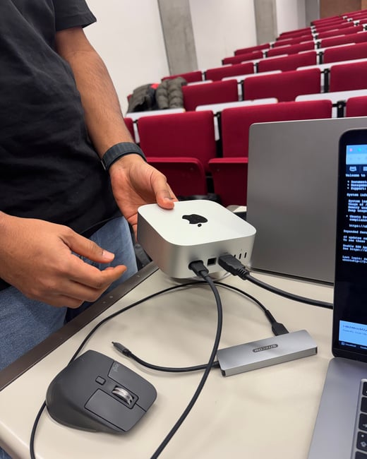 A person is holding an Apple Mac Studio on a desk in a lecture hall, surrounded by computer accessories and a laptop displaying a terminal window.