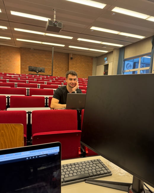A young man sits at a desk in an empty lecture hall with red seats, looking at the camera with a laptop in front of him. A computer monitor and keyboard are positioned in the foreground.