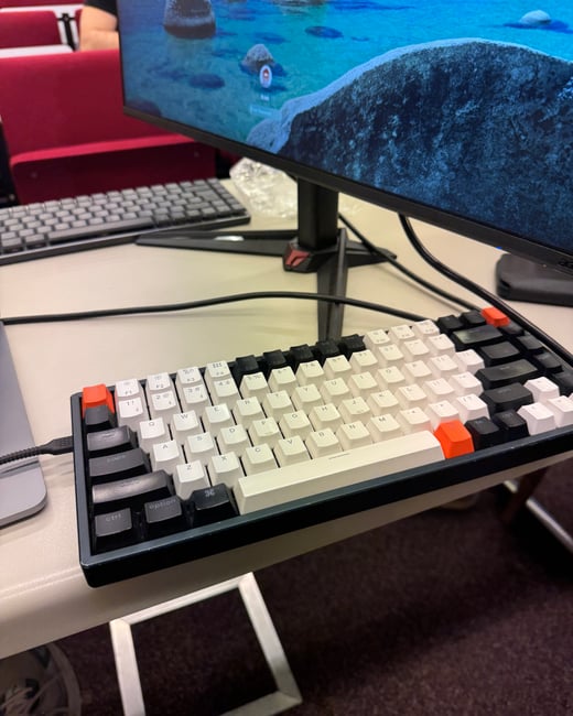 A close-up photograph of a mechanical keyboard with white, black, and orange keycaps on a light-colored desk, with a computer monitor in the background.