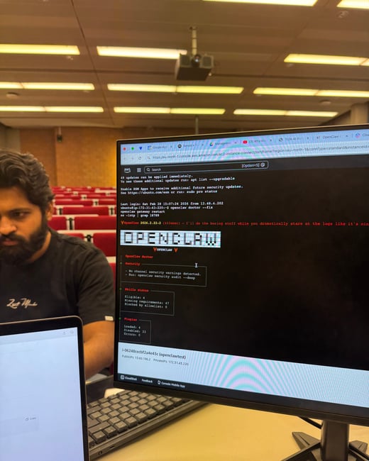 A man sits in an auditorium with red seats, looking down while a computer monitor in the foreground displays a terminal window running 'OpenClaw' software via an AWS console.