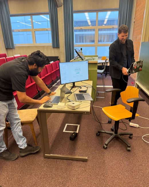 Two men are setting up computer equipment, including multiple laptops and a large monitor, on a desk in a lecture hall with tiered red seating.
