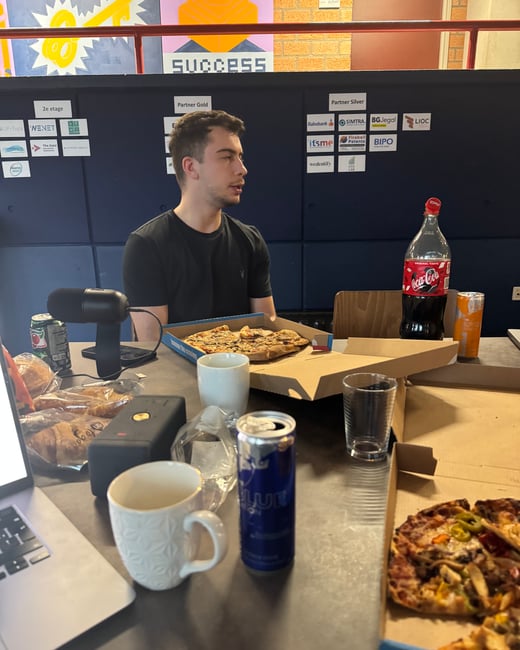 A young man sits at a table cluttered with pizza boxes, various drinks, a laptop, and a microphone, in front of a wall featuring partner logos and the word 'SUCCESS'.