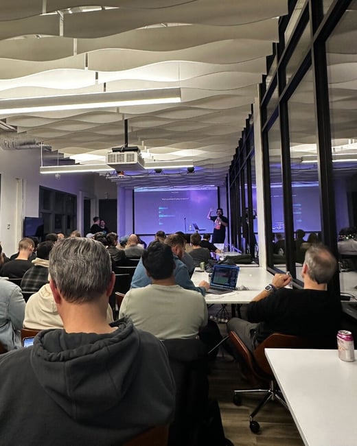 A candid photograph captures a large group of people attending a presentation in a modern office space, featuring a speaker on a stage in front of a projected screen displaying technical content.