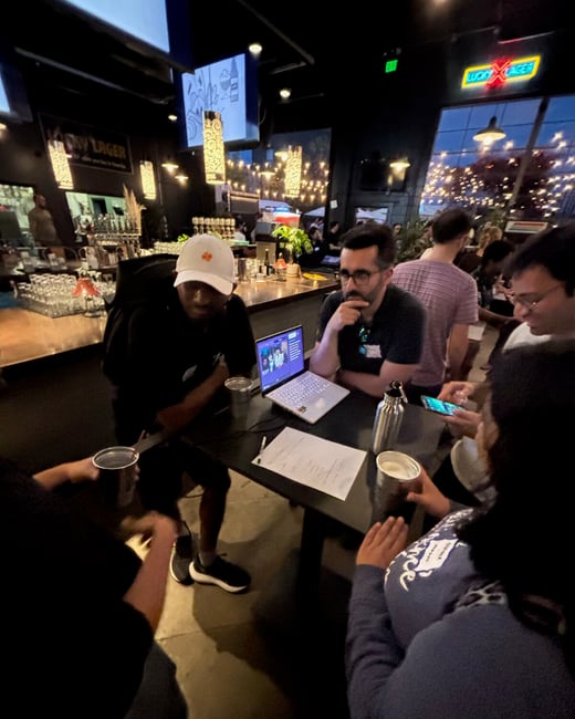 A candid photograph taken inside a dimly lit bar showing a small group of people gathered around a table with an open laptop, seemingly engaged in a discussion or viewing content together.