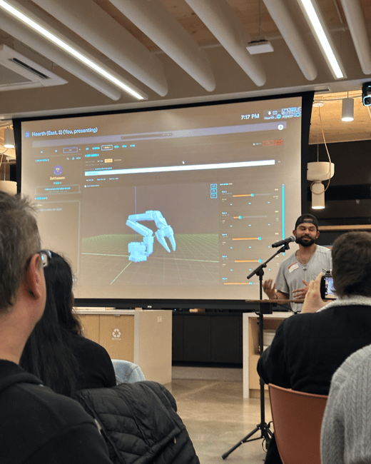 A photograph captures an indoor presentation where a man is speaking into a microphone in front of a large projection screen displaying a 3D simulation of a robotic arm to a seated audience.
