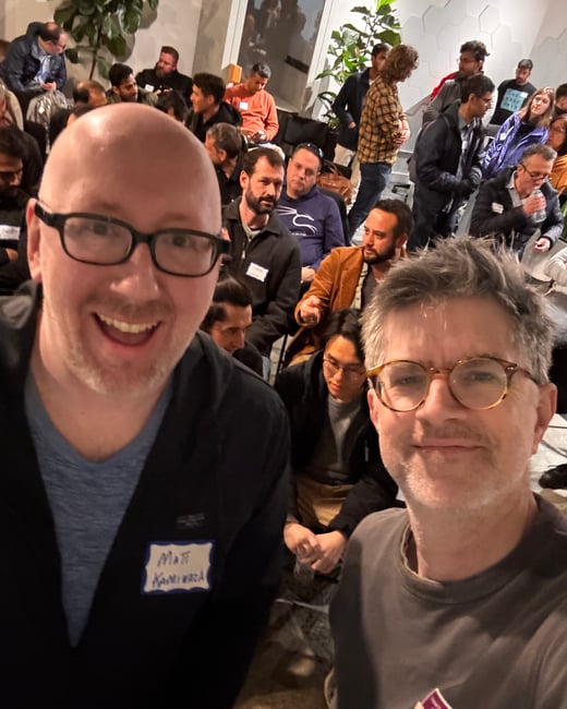 A selfie taken by two men wearing glasses in the foreground, capturing a large, densely packed crowd of attendees behind them at an indoor event or presentation.