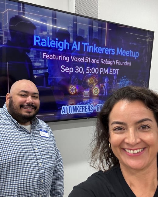 A close-up photograph of a woman and a man smiling at the camera, taken indoors at an event or meeting space. A large screen behind them advertises the 'Raleigh AI Tinkerers Meetup' event.