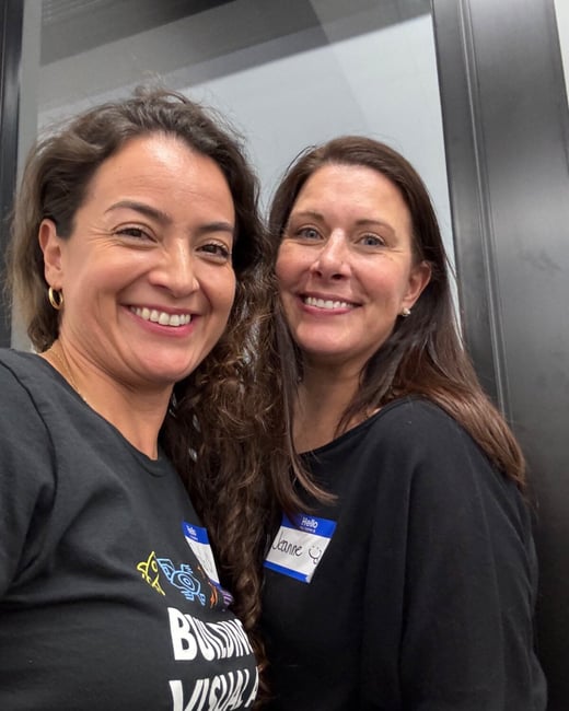 A close-up selfie shows two smiling women standing side-by-side indoors, likely at an event, with one woman wearing a graphic t-shirt and the other wearing a 'Hello my name is' name tag labeled 'Jeanne'.