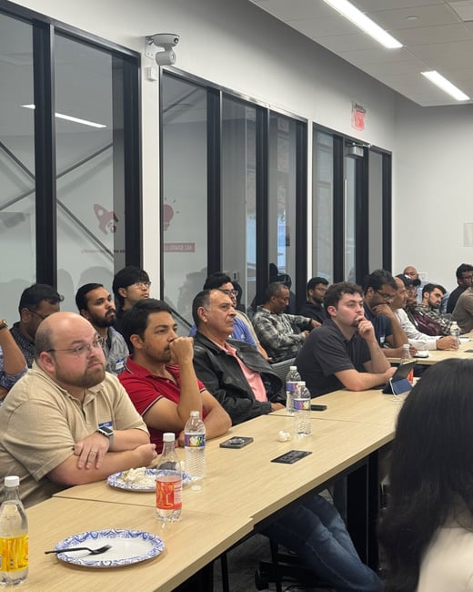 A photograph capturing a group of approximately twenty people seated at a long table in a modern conference room, attentively looking forward, likely attending a presentation or meeting.