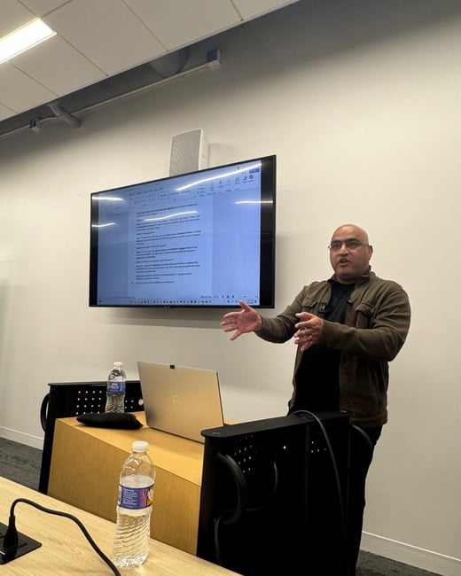 A man is standing and gesturing with his hands while giving a presentation in a modern conference room, with a large wall-mounted screen displaying a document behind him.