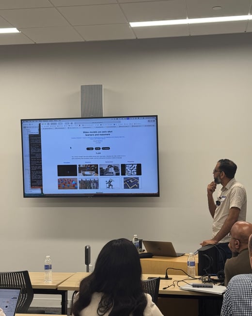 A photograph taken indoors showing a man giving a presentation next to a large wall-mounted screen displaying a slide titled, "Video models are zero-shot learners and reasoners," while several attendees are seated in the foreground.