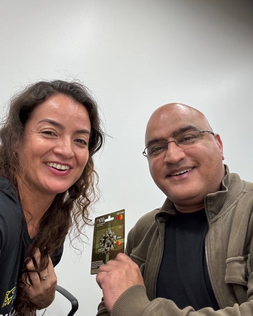 A close-up selfie shows two smiling people, a woman and a man, posing indoors against a plain white wall. The man is holding up a $100 Mastercard gift card towards the camera.