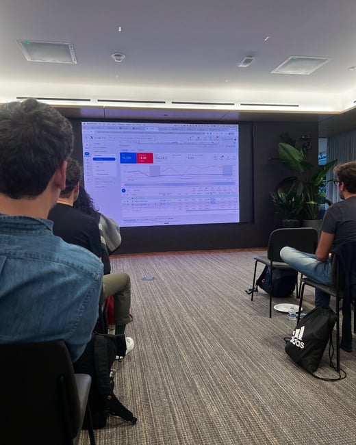 An indoor photograph showing an audience seated in a presentation room, looking at a large screen displaying a Google Ads campaign dashboard.