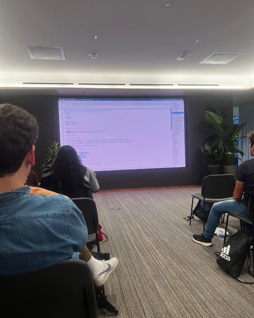 An indoor photograph of an audience viewed from behind, sitting in chairs and watching a presentation displayed on a large screen showing code.