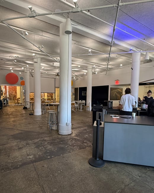 A wide-angle photograph of a spacious, modern indoor area with high ceilings, white pillars, and industrial-style exposed piping. Several people are visible in the background near a counter area.