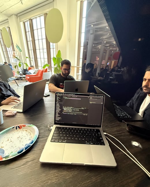 A group of people are working on laptops at a dark wooden table in a bright, modern office space, with one laptop in the foreground displaying code.