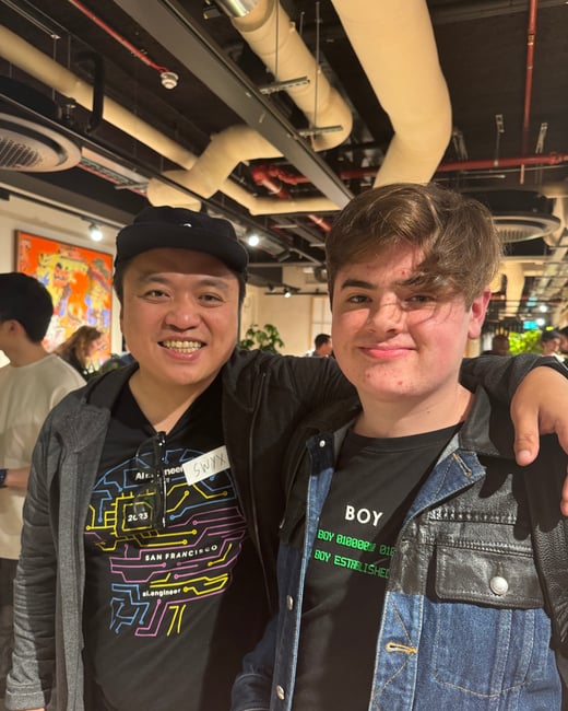 A photo showing two smiling males posing closely together indoors at a casual event or gathering with an exposed industrial ceiling. The man on the left is wearing an event badge and a shirt featuring circuit board graphics.